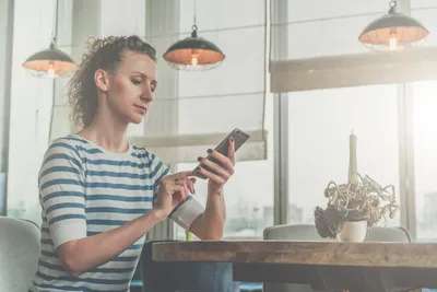 Side view,young serious woman in striped T-shirt sitting in cafe at wooden table in front of window and using smartphone